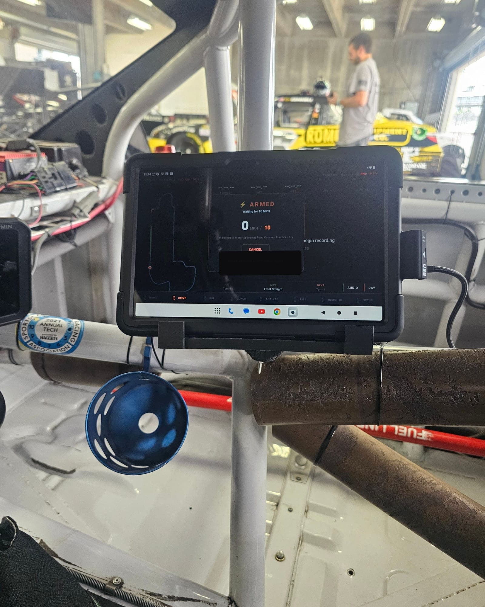 Mission Control Pad mounted on the roll cage of a race car in the Brickyard garage at Indianapolis Motor Speedway, its screen armed with the road course map, while co-founder Nick works on a car in the background